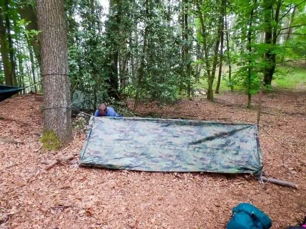 Tarp-Notunterkunft ist zwischen Bäumen gespannt, Person richtet sie ein