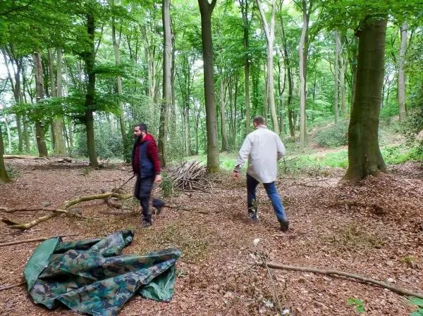 Zwei Personen gehen durch einen Wald, eine Tarp-Notunterkunft liegt am Boden