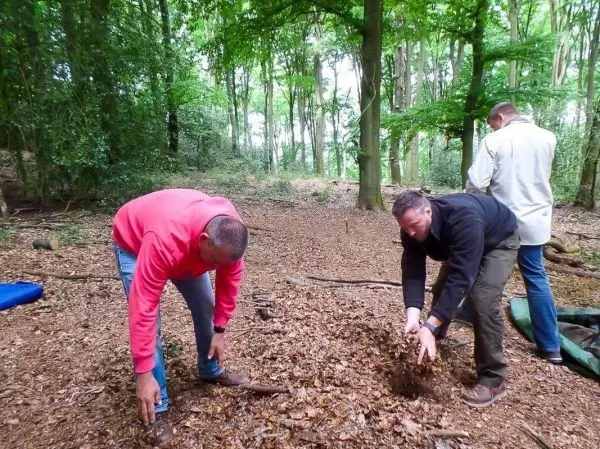 Zwei Personen graben im Waldboden, während eine dritte Person im Hintergrund steht