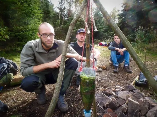 Drei Personen sitzen vor einer improvisierten Wasserfilterkonstruktion aus Holz und Plastikflasche