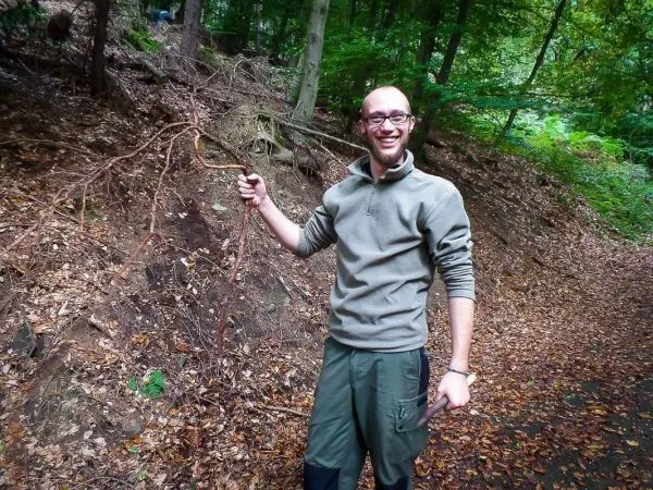 Mann hält einen langen Stock in der Hand, steht auf einem mit Laub bedeckten Weg