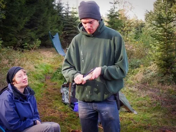 Mann schnitzt mit einem Messer an einem Holzstück, Frau beobachtet