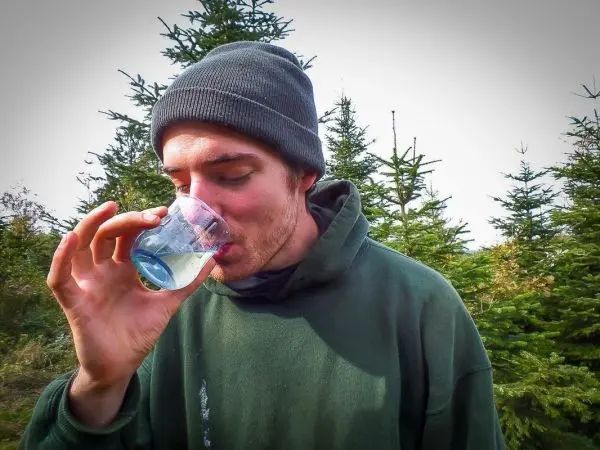 Mann trinkt aus einem Becher inmitten von Tannenbäumen