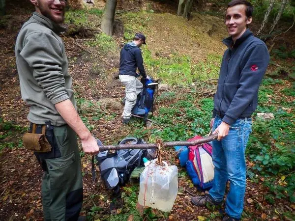 Zwei Personen halten einen Stock mit einem Behälter, während eine dritte Person Rucksäcke organisiert