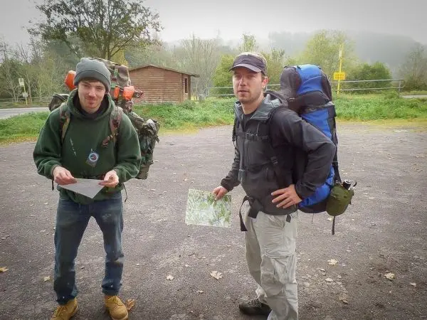 Zwei Personen mit Rucksäcken studieren Kartenmaterial auf einem Parkplatz