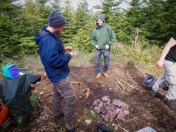Zwei Personen stehen in einem Waldgebiet neben einem Steinhaufen für ein Feuer