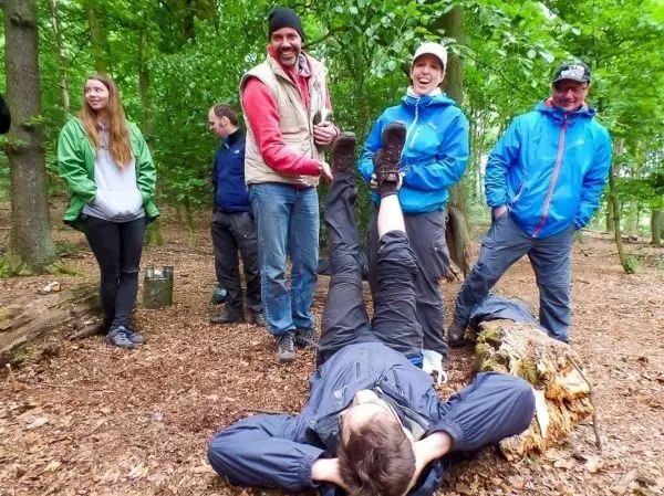 Gruppe von Personen steht im Wald, eine Person liegt auf dem Boden und hebt die Beine