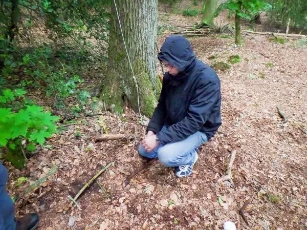 Mann kniet neben einer selbstgebauten Schlagfalle unter einem Baum