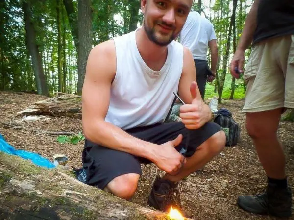Kleines Bodenfeuer brennt neben einem Holzstamm, Person gibt Daumen hoch