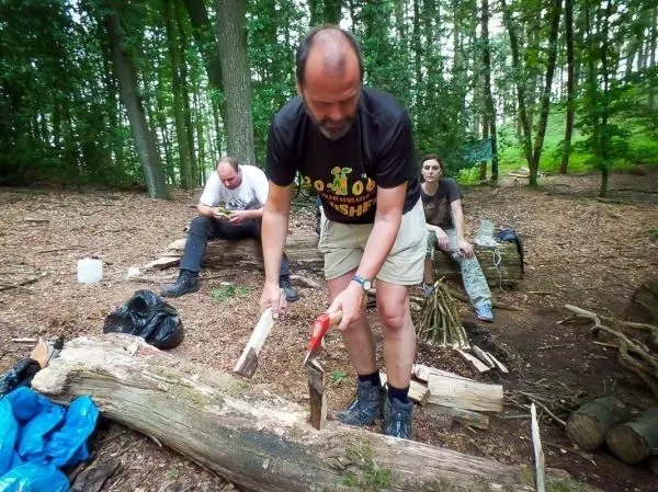 Mann bearbeitet Holzstück mit Axt, im Hintergrund liegen weitere Holzstücke