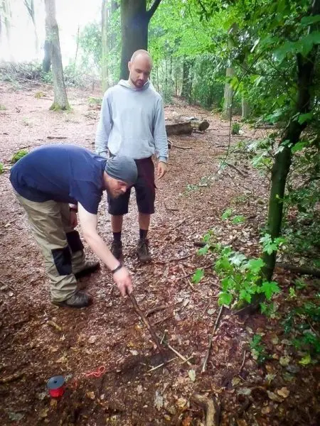 Mann gräbt mit einem Stock im Waldboden, während ein anderer zuschaut