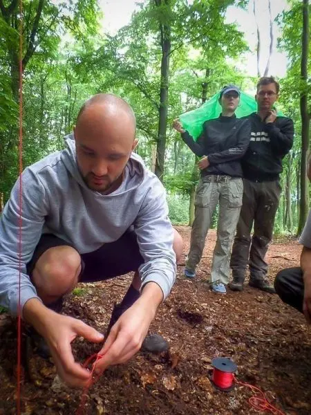 Mann kniet im Wald und knotet ein rotes Seil