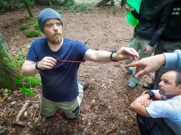 Mann kniet im Wald und zeigt auf eine Schnur, während andere zuschauen