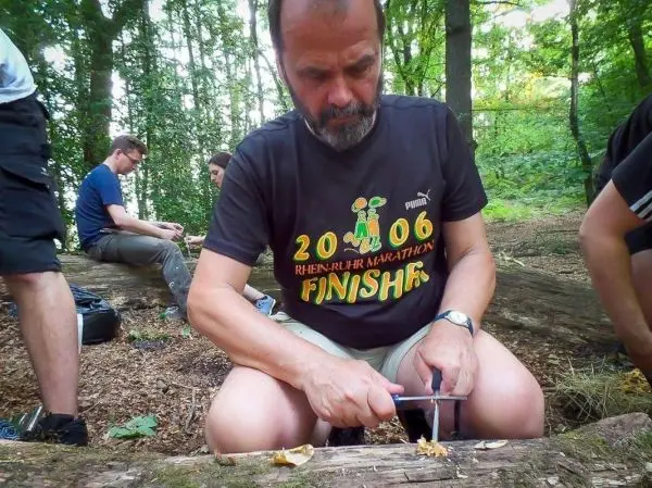 Mann schnitzt mit einem Messer an einem Holzstück im Wald
