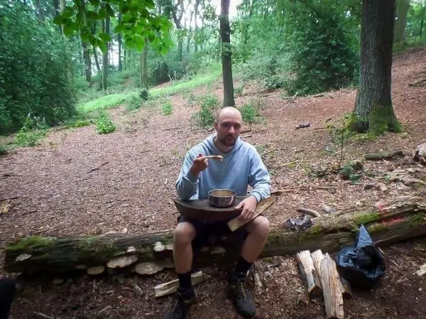 Mann sitzt auf einem Baumstamm und hält einen Löffel über einem Kochtopf