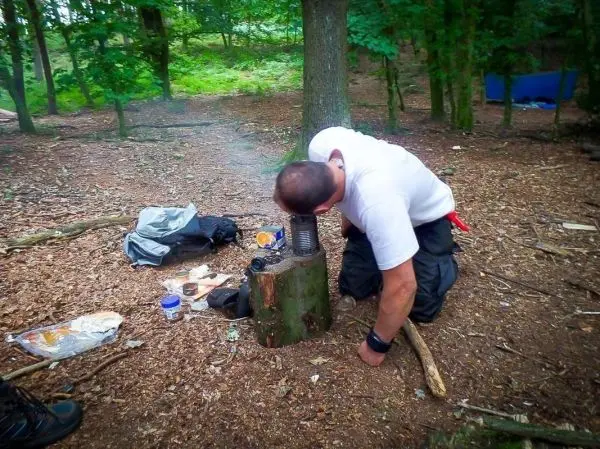 Person beugt sich über einen Hobo-Kocher auf einem Baumstumpf im Wald