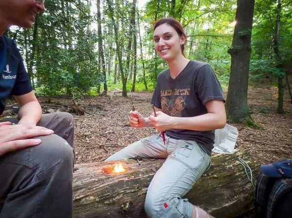 Person sitzt auf einem Baumstamm und hält einen Stock über Feuer beim Survival Training