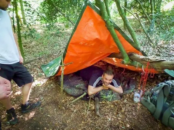 Tarp-Notunterkunft aus orangefarbener Plane und Ästen, Person liegt darunter