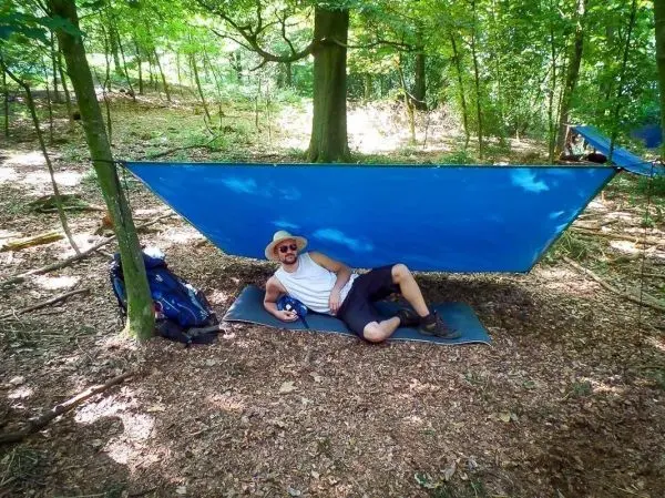 Tarp-Notunterkunft hängt zwischen Bäumen, Person liegt entspannt darunter auf einer Matte