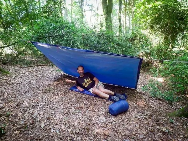 Tarp-Notunterkunft ist zwischen Bäumen gespannt, Person liegt auf einer Matte darunter