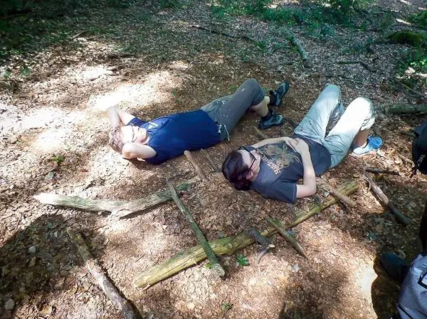Zwei Personen liegen auf einem improvisierten Untergrund aus Ästen im Wald