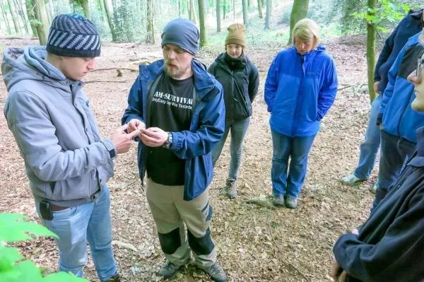 Ein Mann zeigt einem anderen eine Technik im Wald, während mehrere Personen zuschauen