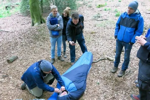 Eine Person in einem Schlafsack erhält Erste Hilfe von einer anderen Person im Wald
