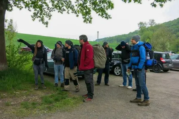 Gruppe mit Rucksäcken steht an einem Parkplatz und hört einer Person zu