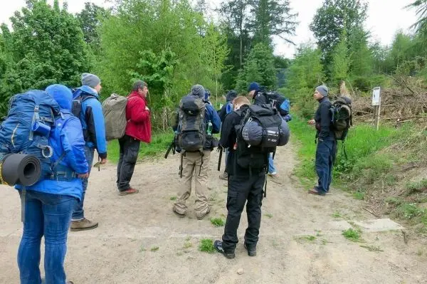 Gruppe mit Rucksäcken steht an einem Waldweg und hört einem Vortrag zu