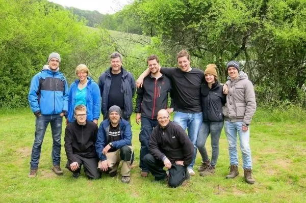 Gruppe von Personen steht und sitzt auf einer Wiese umgeben von Bäumen