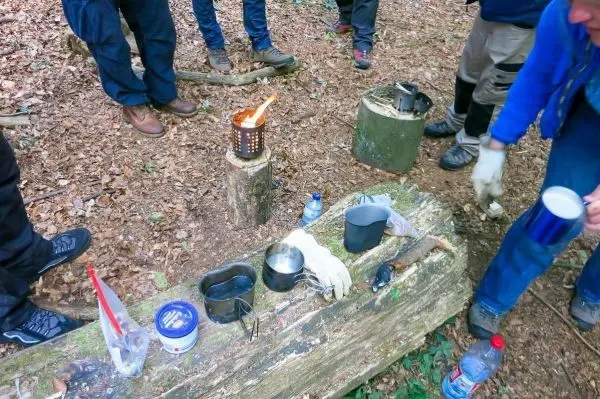 Kessel auf Holzstamm, daneben Spirituskocher und Wasserflasche, mehrere Personen umgeben die Szene