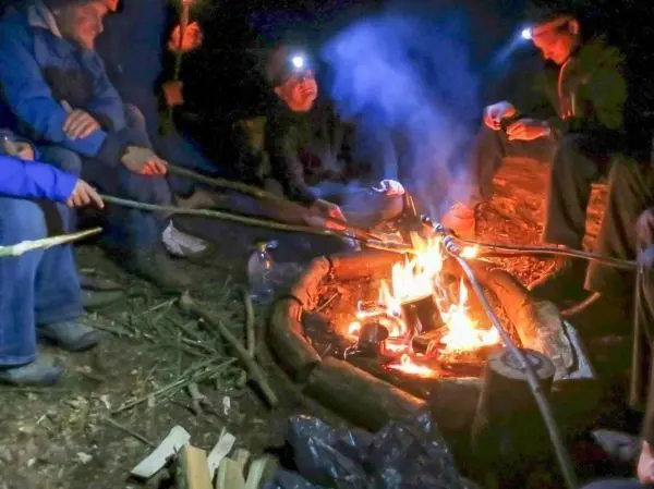 Personen sitzen um ein Feuer und halten Stöcke in der Hand beim Überlebenstraining