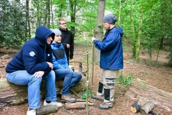 Vier Personen sitzen und stehen um einen Baumstamm im Wald, einer erklärt eine Technik mit einem Stock
