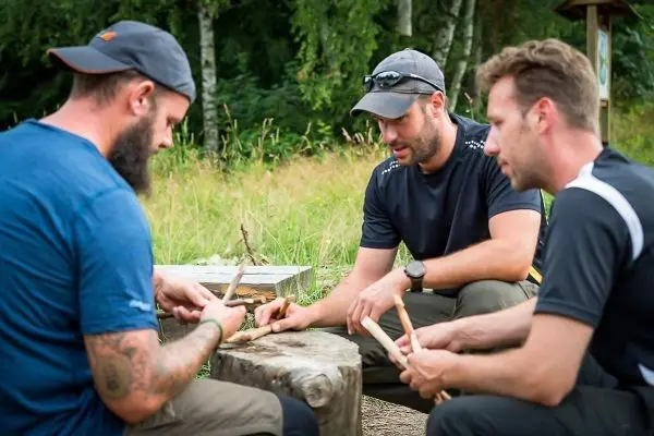Drei Personen bearbeiten Holzstücke auf einem Baumstamm im Freien