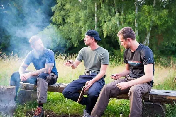 Drei Personen sitzen auf einer Holzbank und unterhalten sich am Lagerfeuer
