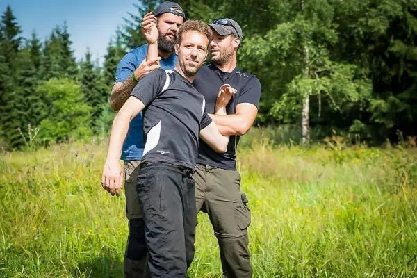 Drei Personen stehen in einer Wiese, eine Person wird von den anderen umarmt