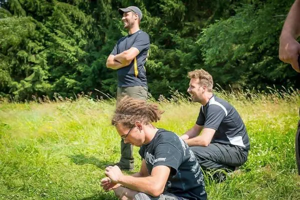 Drei Personen stehen und sitzen im Gras, eine Person bearbeitet etwas mit den Händen