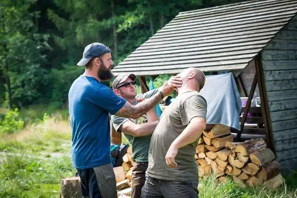 Drei Personen üben eine Selbstverteidigungstechnik in der Nähe einer Holzunterkunft