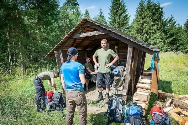 Holzhütte im Wald mit mehreren Rucksäcken und Personen, die Ausrüstung überprüfen