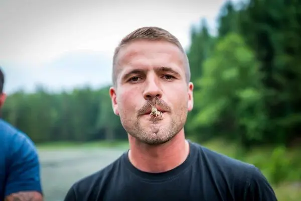 Mann mit Insekt im Mund steht vor bewaldetem Hintergrund