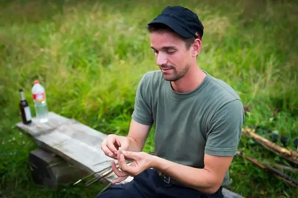 Mann sitzt auf Holzbank und bearbeitet einen Stock mit einem Messer