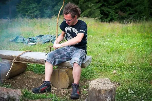 Mann sitzt auf Holzbank und bearbeitet einen Stock mit einem Messer