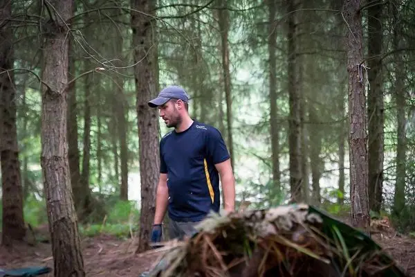 Mann steht in einem Wald neben einer improvisierten Unterkunft aus Ästen und Blättern