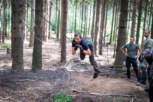 Mann überquert selbstgebaute Seilbrücke im Wald, Zuschauer beobachten