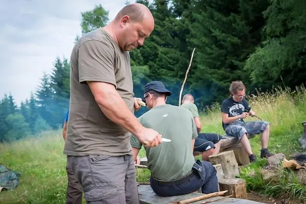 Messer bearbeitet Holzstück auf einem Baumstamm, im Hintergrund weitere Personen