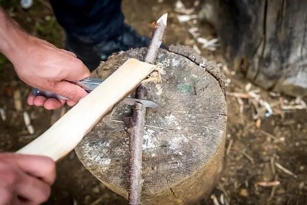 Person schnitzt mit einem Messer einen Stock auf einem Baumstumpf