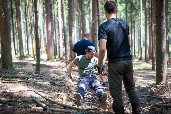 Person sitzt auf dem Boden zwischen Ästen, zwei Personen stehen daneben
