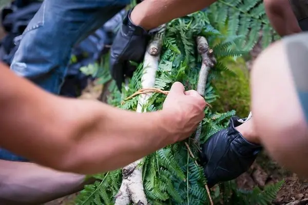 Zwei Hände binden Äste mit Pflanzenmaterial zu einer improvisierten Trage