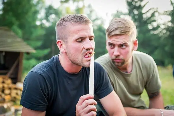 Zwei Männer sitzen im Freien, einer hält einen langen Stock und schaut konzentriert