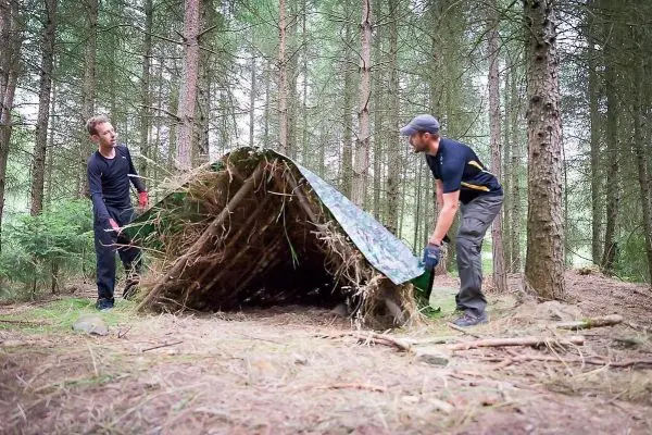 Zwei Personen bauen eine Tarp-Notunterkunft aus Ästen und einer Plane auf